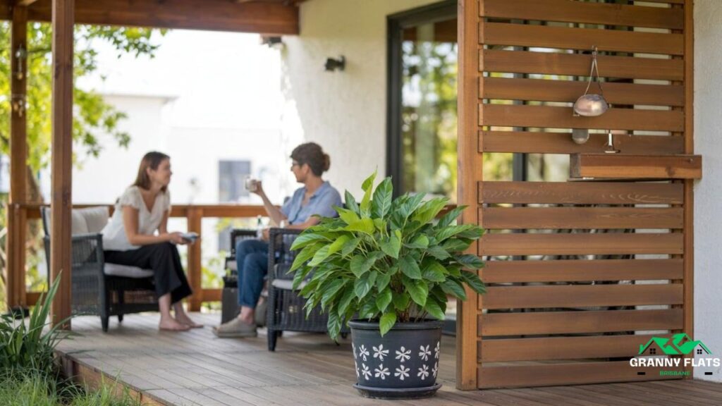 Two women chatting on a timber granny flat deck with potted plants and outdoor seating.